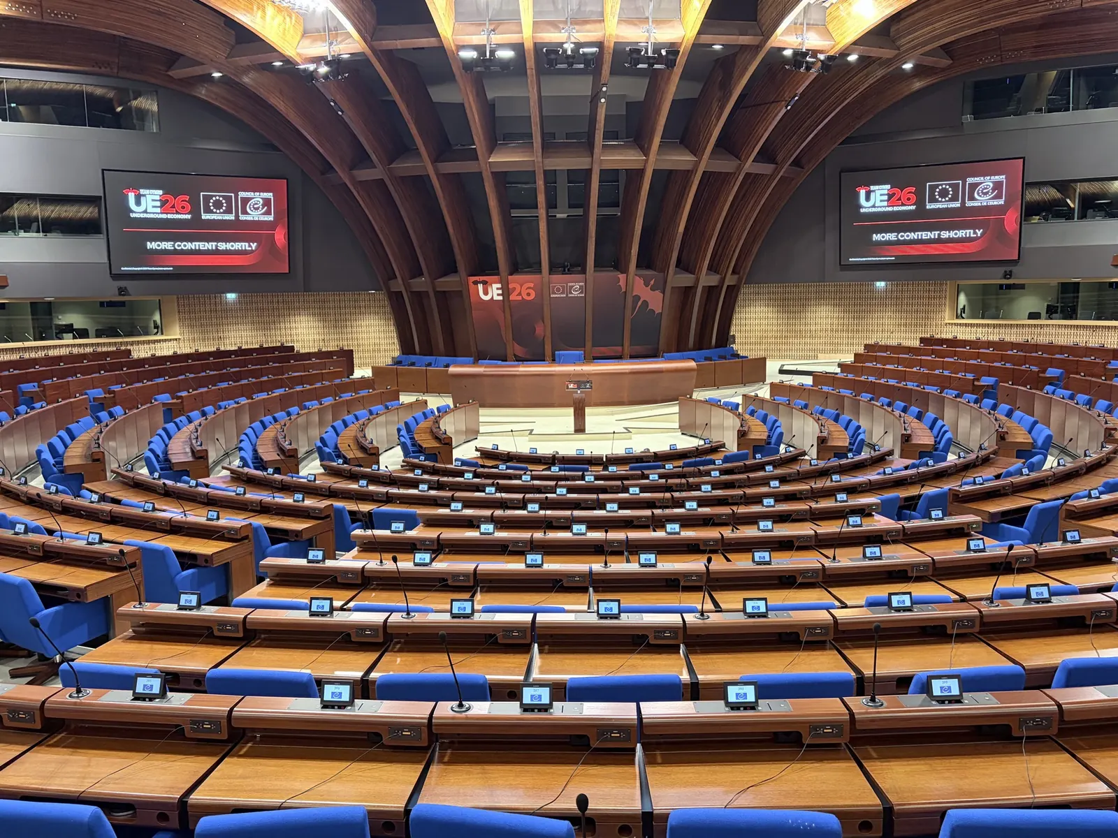 The Council of Europe hemicycle in Strasbourg, empty before a session, its curved rows of blue seats and wooden desks facing a podium. Screens either side carry the UE26 Underground Economy logo alongside the European Union and Council of Europe marks.